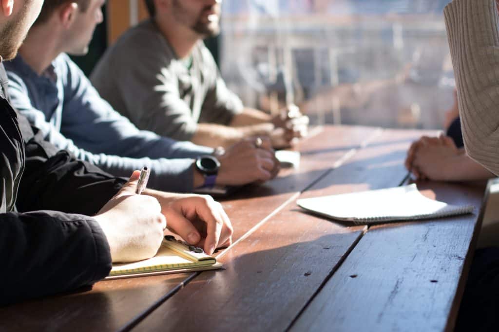 Close-up of people at a meeting at a wooden table. They are holding pens and notebooks.